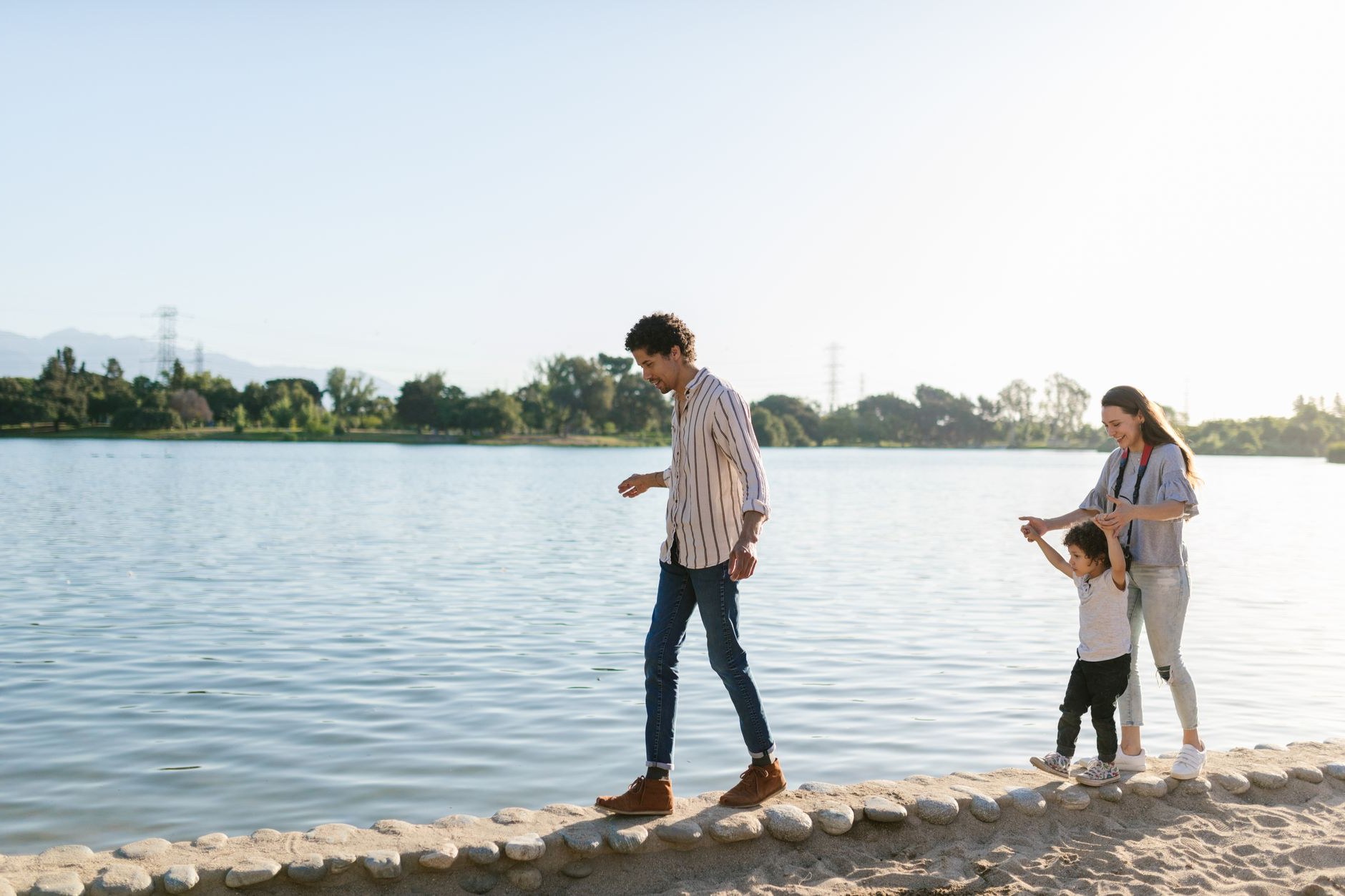 A parent holding a child&rsquo;s hand while walking beside a calm lake